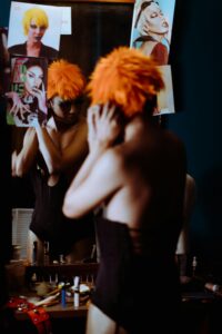 My Personal Gay Stories Back view bizarre female in black bodysuit putting on bright orange wig against mirror in dressing room