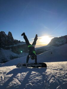 Queer Destinations Snowboarder performs a handstand on a snowy slope at sunset, surrounded by mountains.