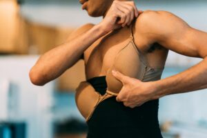 Crop anonymous shirtless drag queen putting on bra while standing in dressing room on blurred background